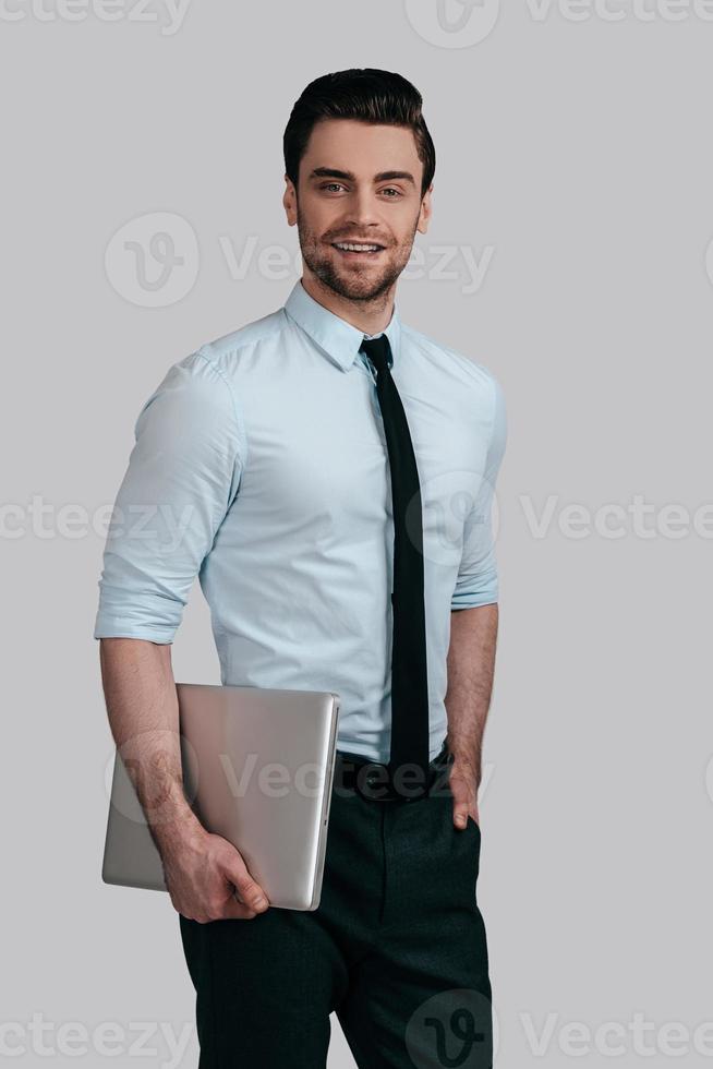 Confident businessman. Good looking young man in formalwear carrying laptop and looking at camera while standing against grey background photo