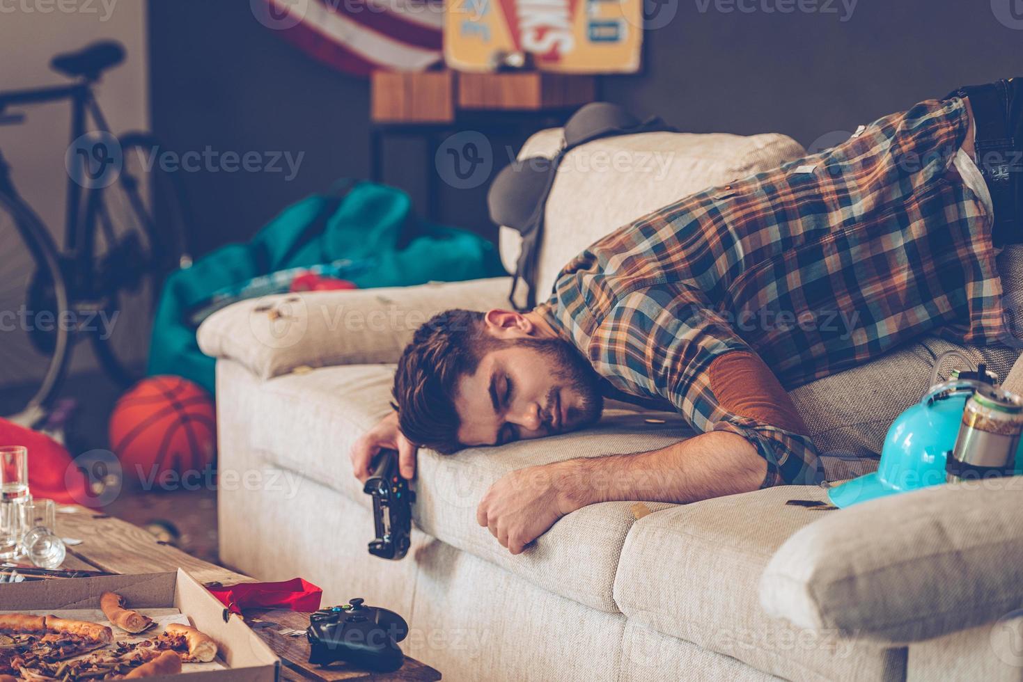 Party is over. Young handsome man passed out on sofa with joystick in his hand in messy room after party photo