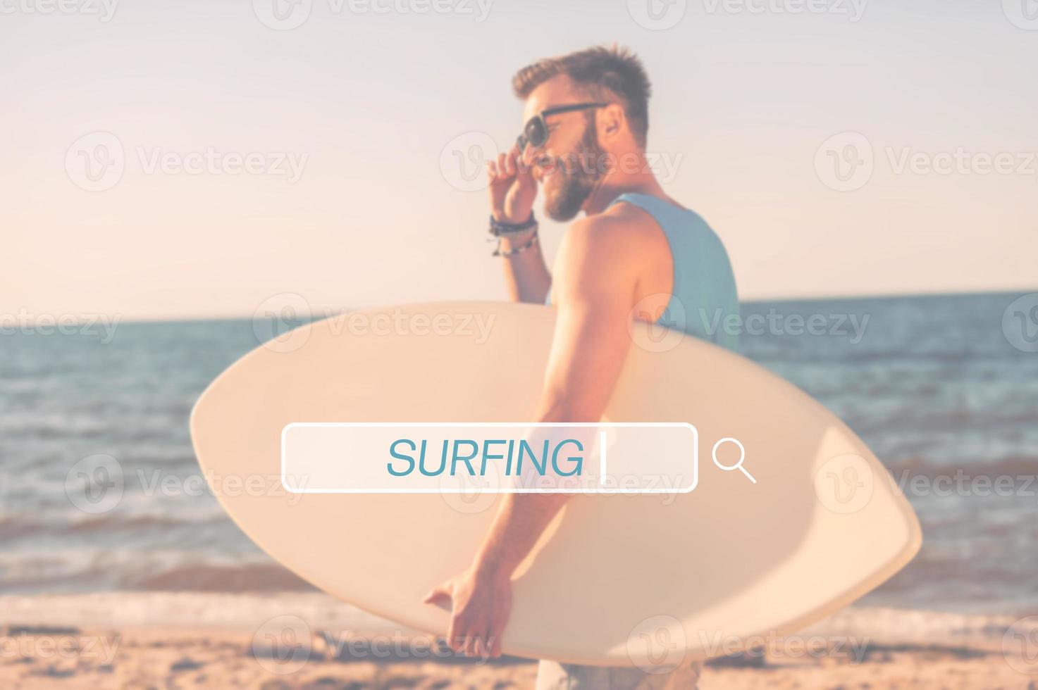 Find your place for surfing. Handsome young man holding skimboard and adjusting eyewear while walking along the beach photo