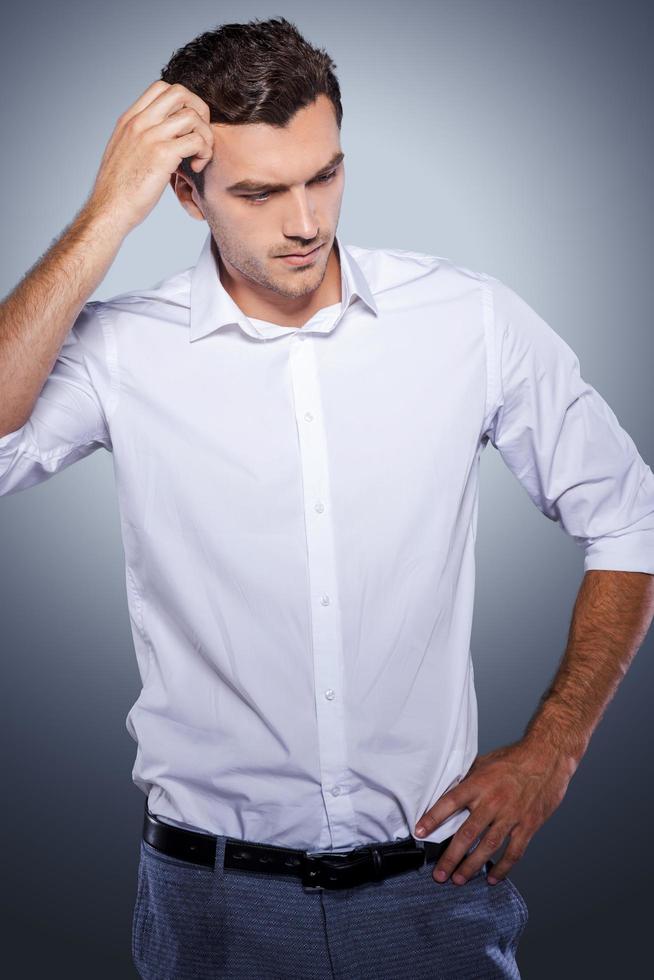 Lost in thoughts. Handsome young man in white shirt holding hand in hair and looking away while standing against grey background photo