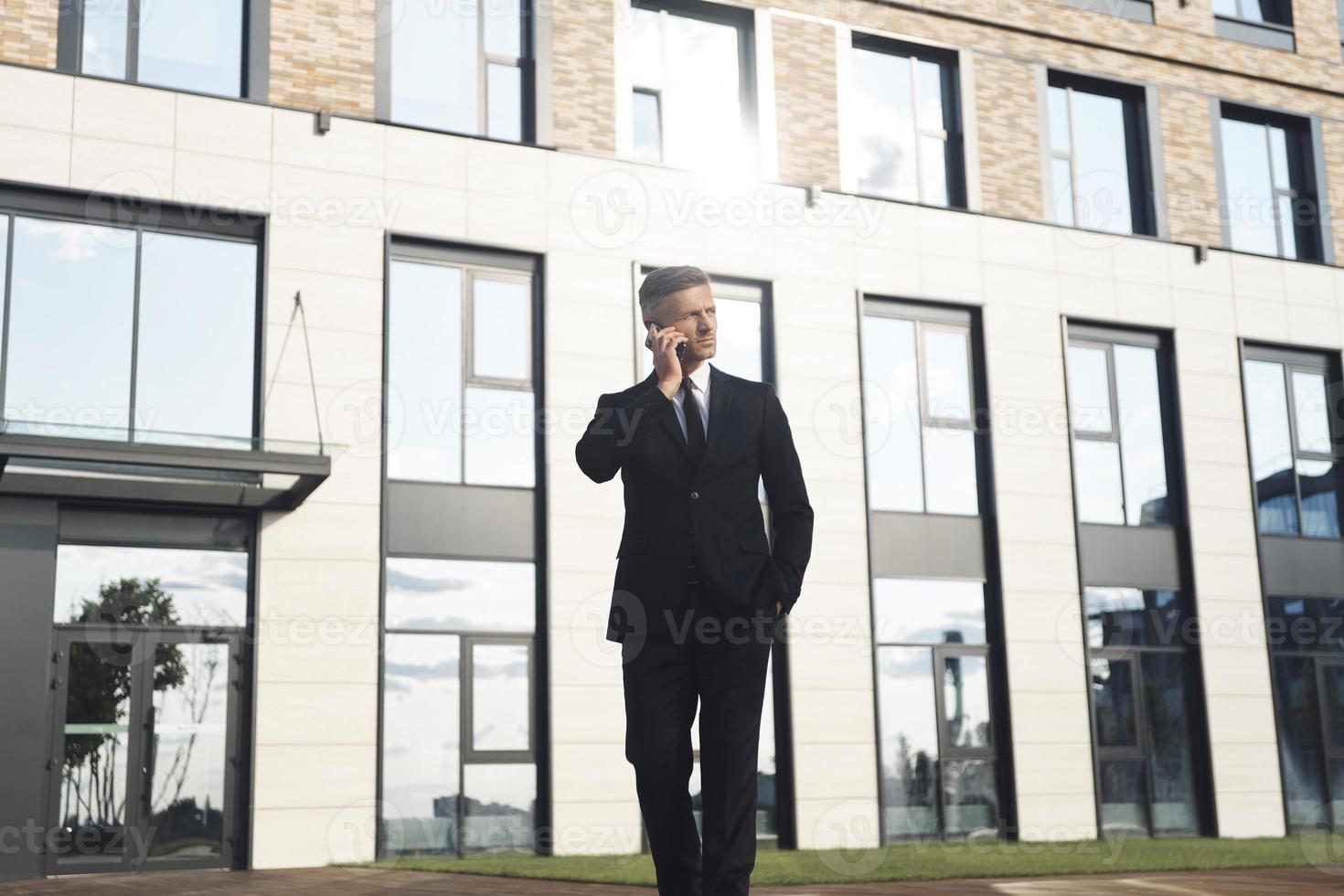 Confident mature businessman talking on mobile phone while walking in front of office building photo