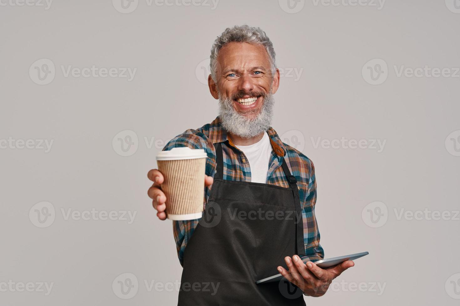 Senior man in apron smiling and giving you coffee while standing against gray background photo