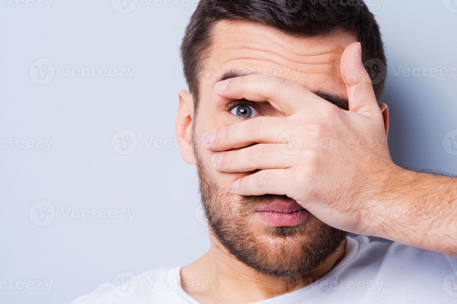 Shocked and terrified. Portrait of young man covering his face by hand and looking at camera while standing against grey background photo