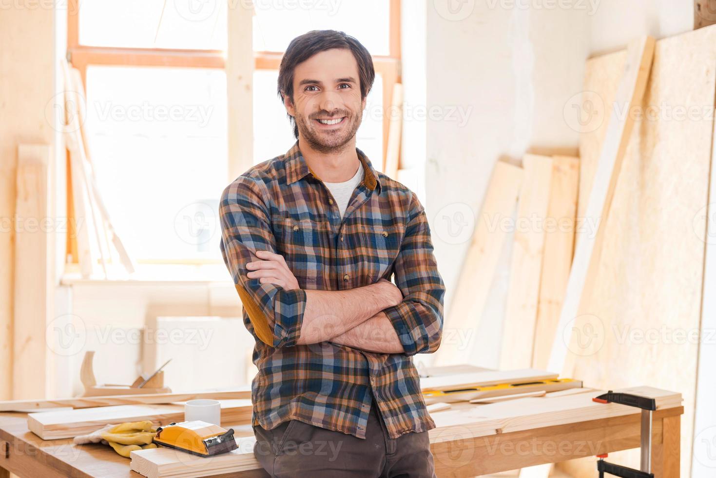 My workshop is my world Happy young male carpenter keeping arms crossed and leaning at the wooden table with diverse working tools laying on it photo