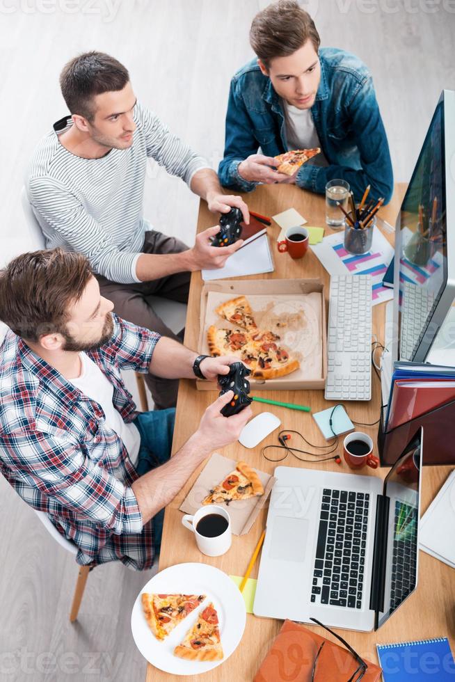 Relaxing after work. Top view of three young men playing computer games and eating pizza while sitting at the desk photo