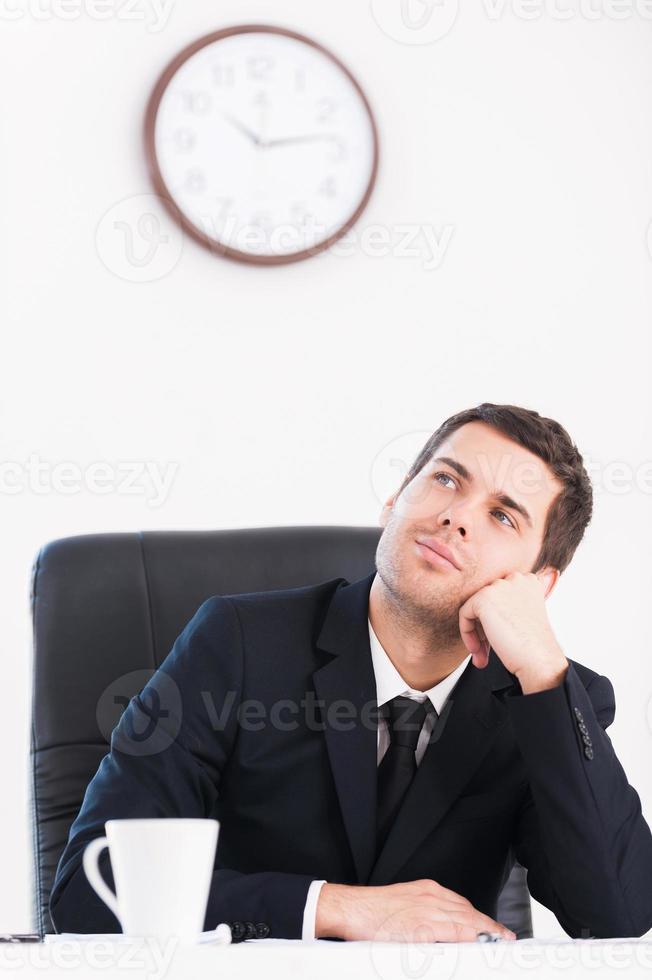 Break time. Thoughtful young man in formalwear looking away and holding head in hand while sitting at his working place with wall clock on background photo