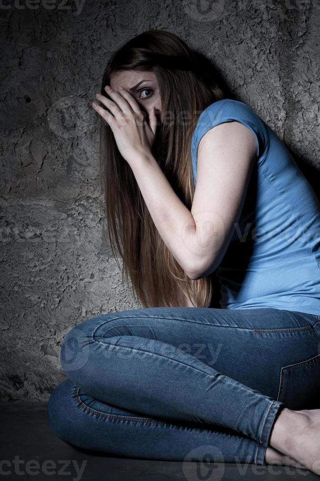 Please no Shocked young woman covering face with hands and looking away while sitting on the floor in a dark room photo
