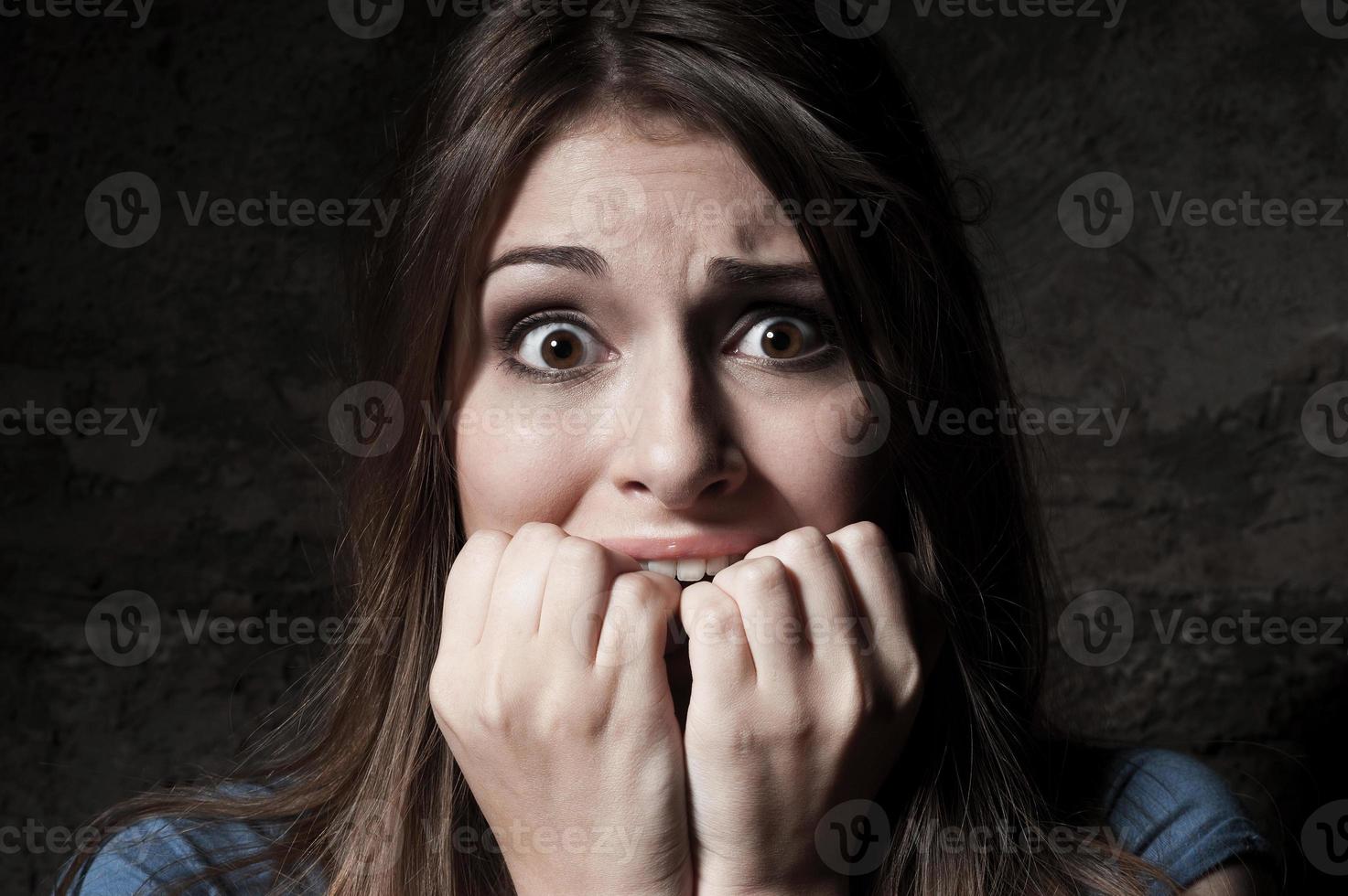I am so scared Shocked young woman staring at camera while holding fingers in mouth while standing against dark background photo