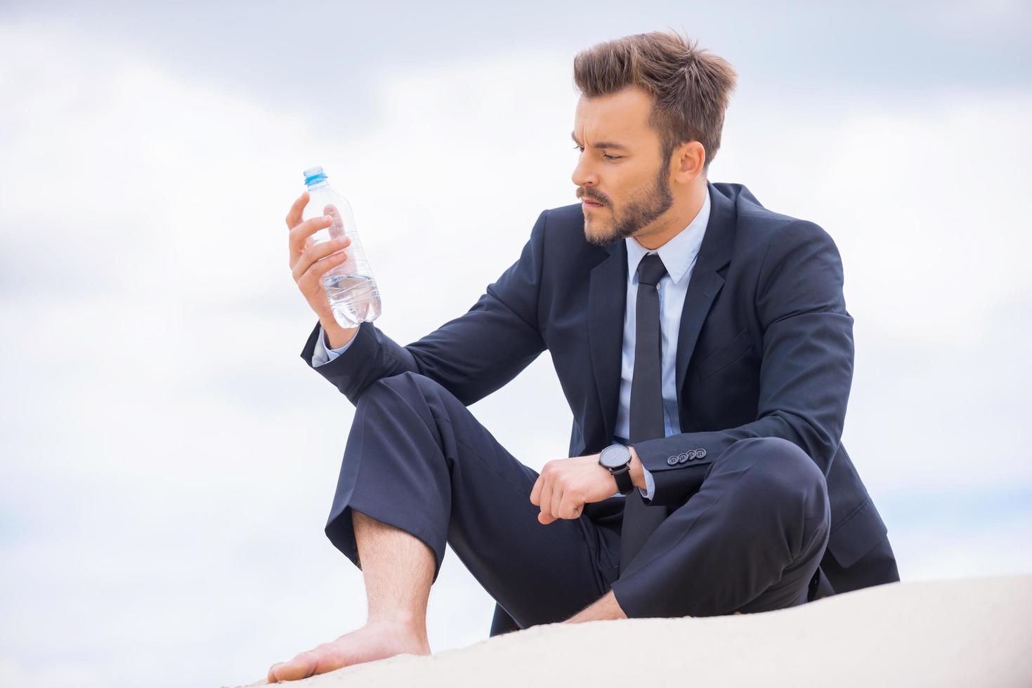 He needs to get refreshed. Depressed young businessman holding bottle with water and looking at it while sitting on sand photo