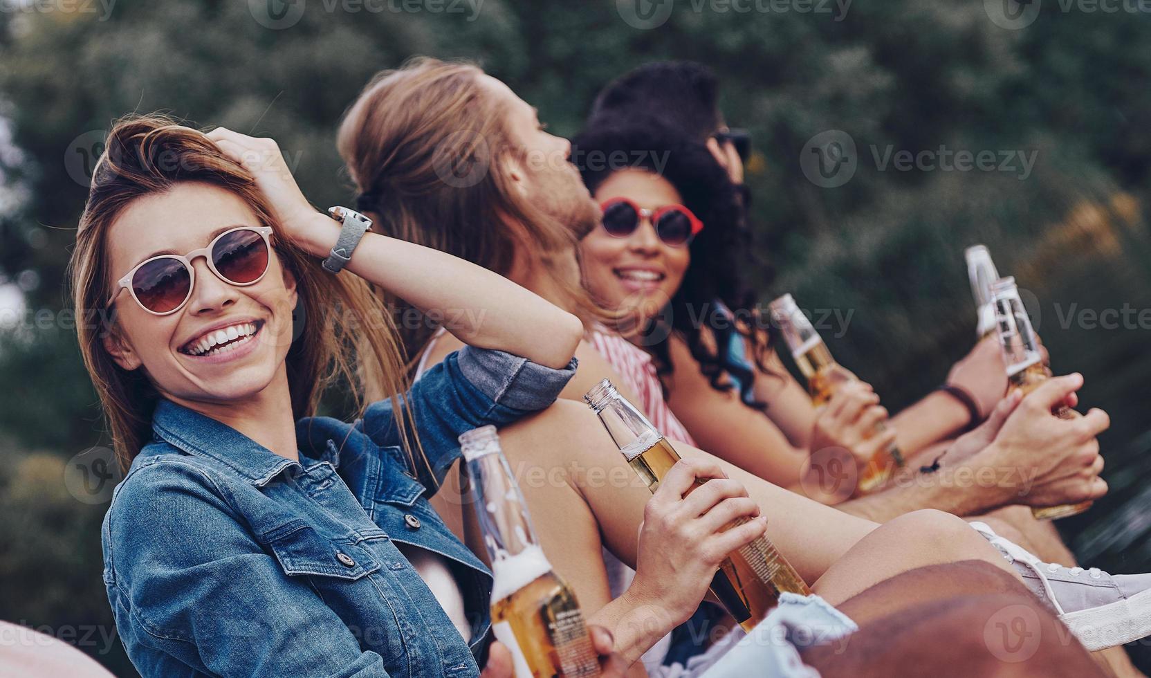 Leave all the worries behind. Group of happy young people in casual wear smiling and drinking beer while sitting on the pier photo