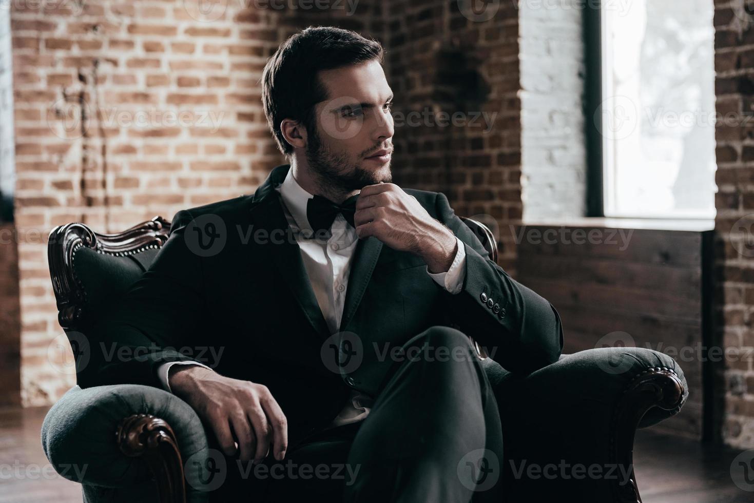 Royalty in his blood. Thoughtful young handsome man in formalwear and bow tie looking away and holding hand on chin while sitting in a chair in loft interior photo