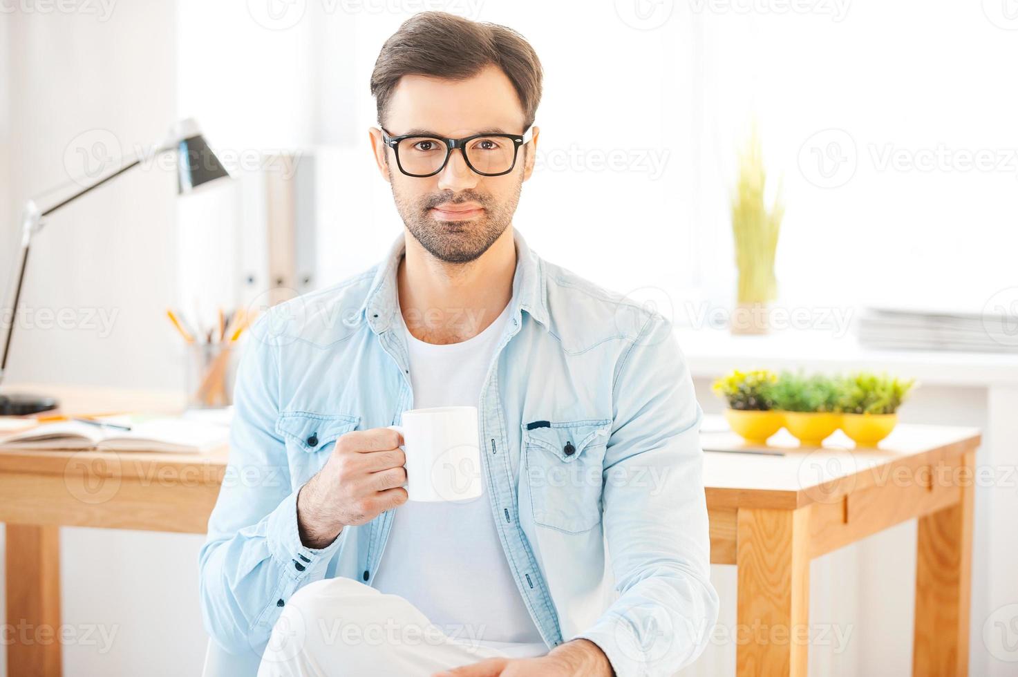 Cup of coffee for great ideas. Handsome young man in shirt and eyewear holding cup of coffee and looking at camera while sitting in front of his working place photo