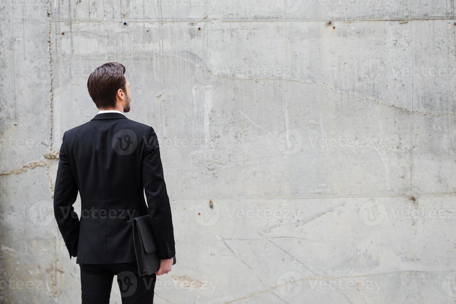 Facing a wall. Rear view of young man holding briefcase while standing outdoors and against the concrete wall photo