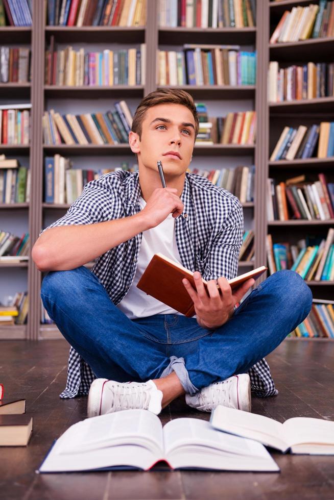 Waiting for inspiration. Thoughtful young man holding note pad and touching his chin with pen while sitting against bookshelf photo