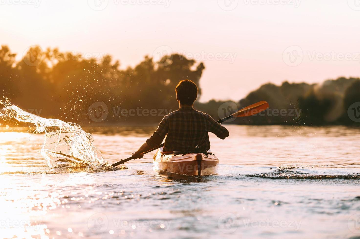 hombre en kayak. vista trasera del joven salpicando agua mientras navega en kayak en el río con la puesta de sol en el fondo foto