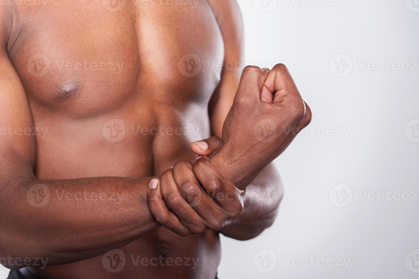 Feeling pain in joint. Cropped image of young muscular African man touching his hand while standing against grey background photo
