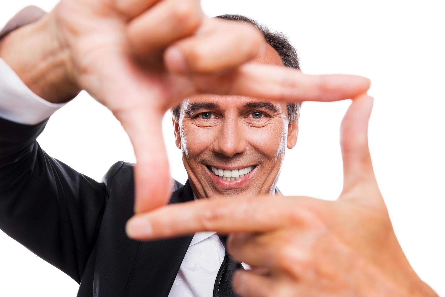 Focusing at you. Cheerful mature man in formalwear gesturing finger frame and looking through it while standing isolated on white background photo