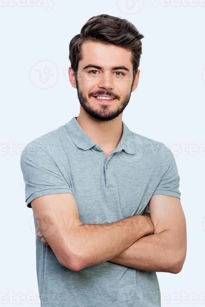 Candid smile. Handsome young man keeping arms crossed and looking at camera while standing against white background photo