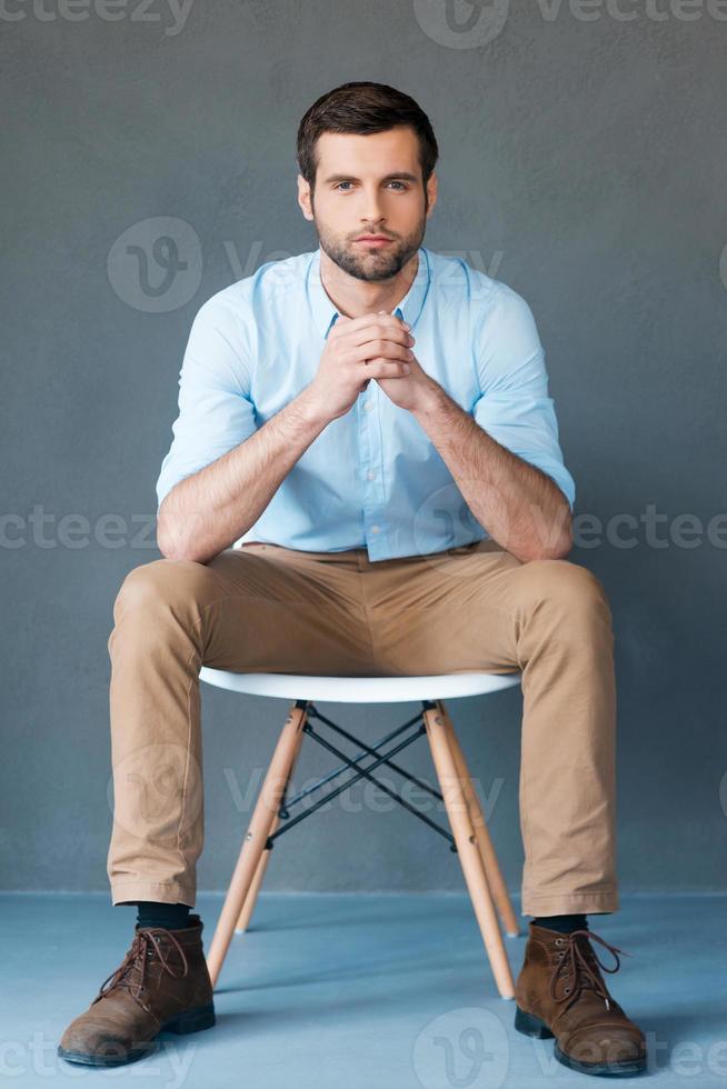 Smart and confident. Full length of handsome young man looking at camera while sitting against grey background photo