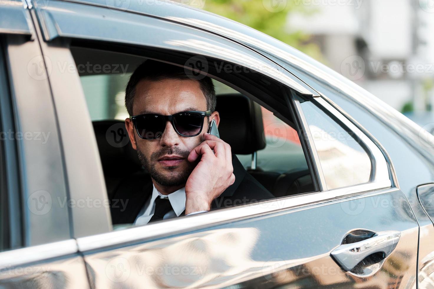 Confident and successful. Serious young businessman talking on the mobile phone while riding inside his car photo