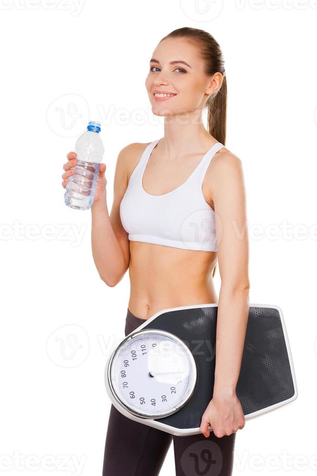 Living a healthy life. Attractive young woman in sports clothing holding weight scale and bottle with water while standing isolated on white photo