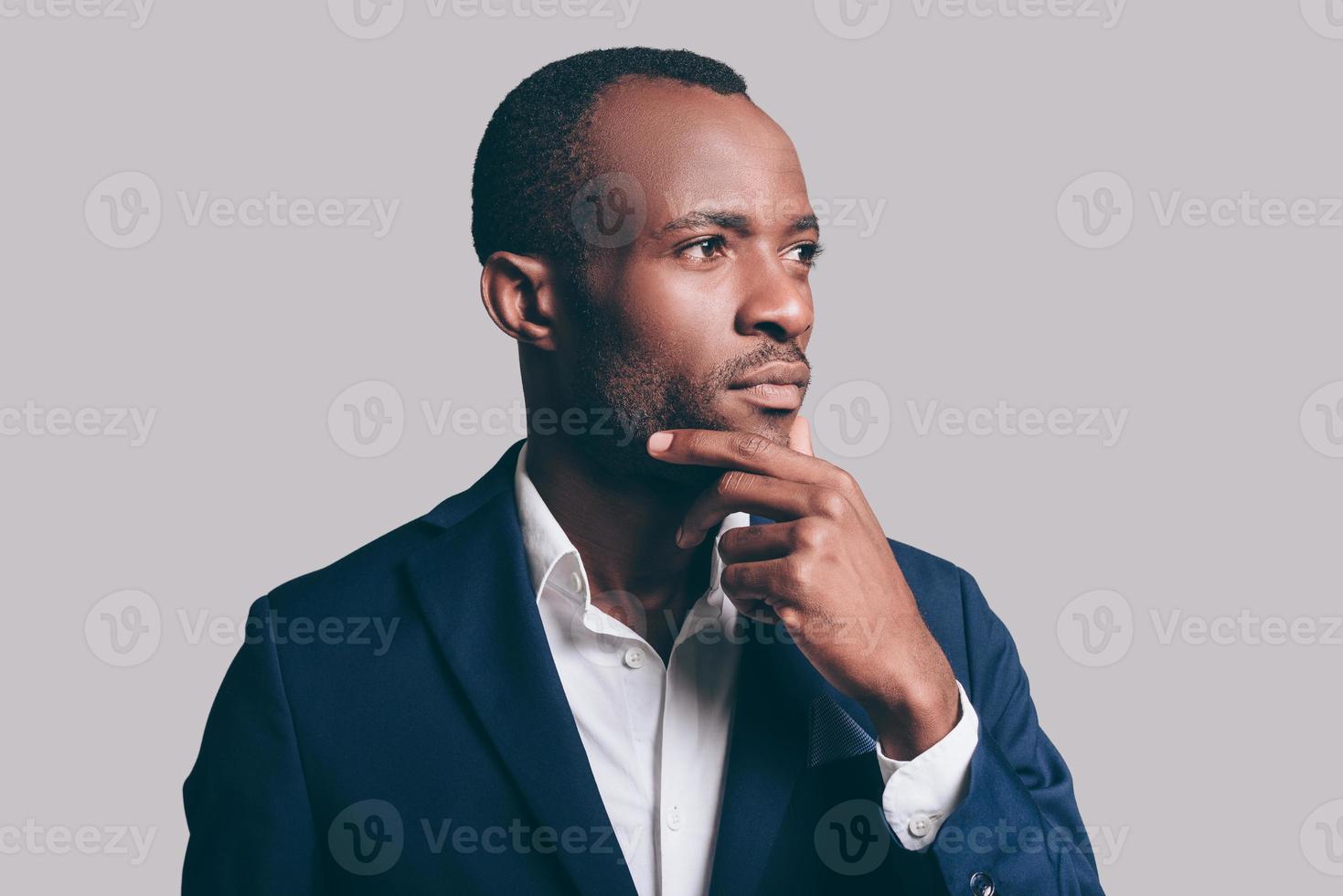 Lost in thoughts. Portrait of thoughtful young African man in smart casual jacket holding hand on chin and looking away while standing against grey background photo