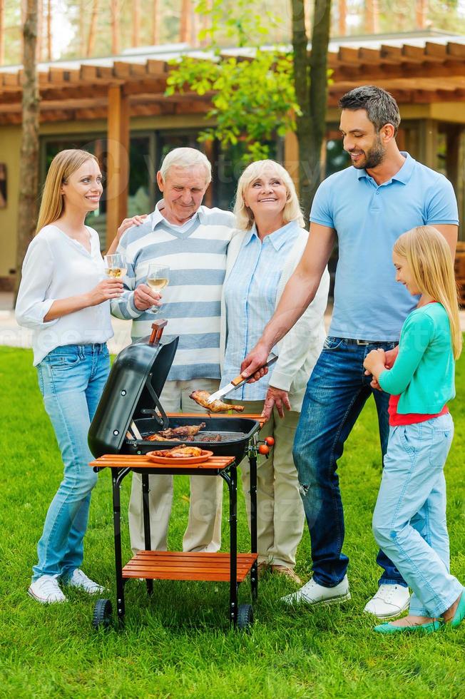 Time for barbeque. Full length of happy family barbecuing meat on grill outdoors photo