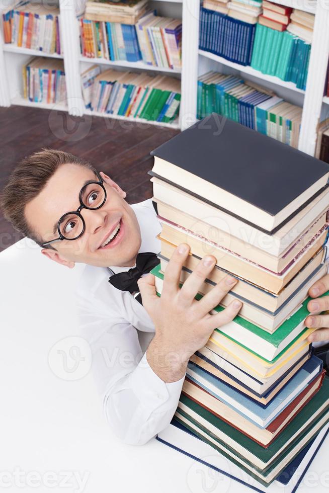 His favorite books. Top view of excited young nerd man in shirt and bow tie sitting at the table in library and hugging a book stack photo