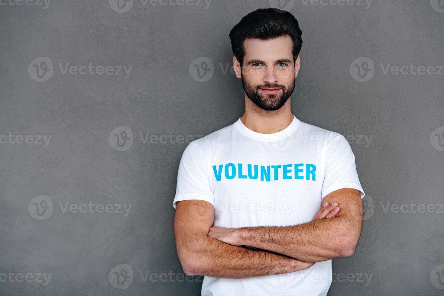 Ready for charity work. Confident young man in volunteer t-shirt keeping arms crossed and looking at camera with smile while standing against grey background photo