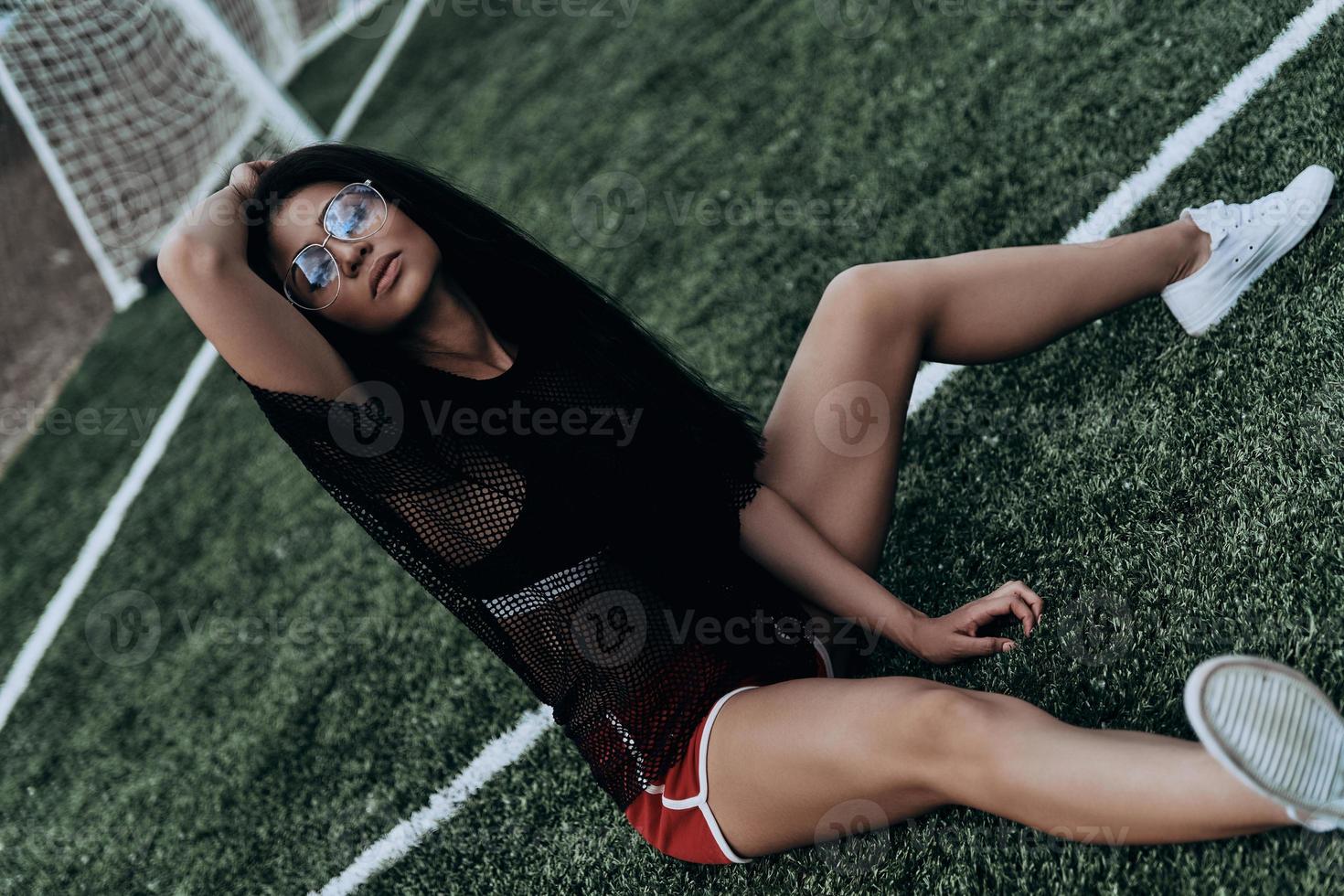 Time to relax. Beautiful young woman in sport clothing keeping hand in hair while sitting on the soccer field photo