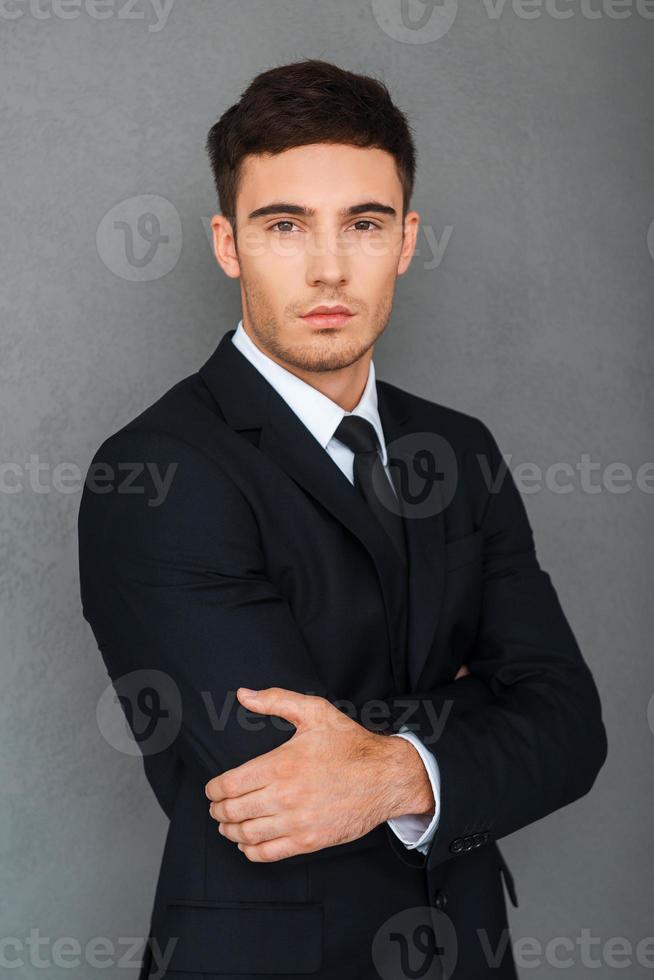 Young and successful. Confident young businessman keeping arms crossed and looking at camera while standing against grey background photo