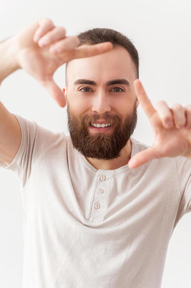 Focusing on you. Handsome young bearded man focusing on you and smiling while standing isolated on grey background photo