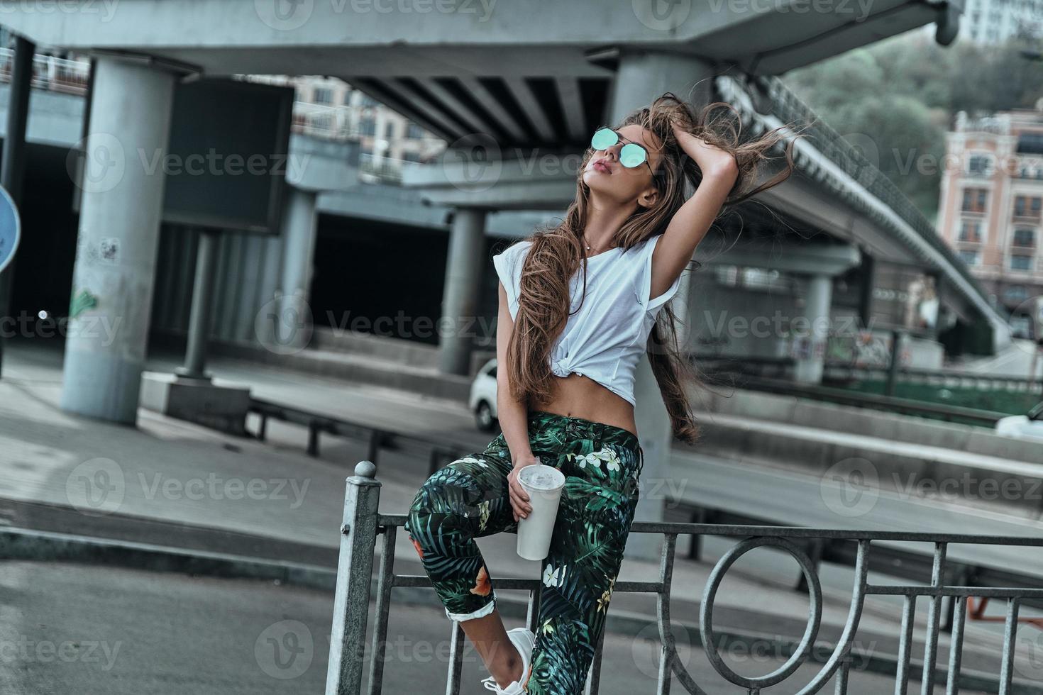 chica de ciudad. hermosa joven manteniendo la mano en el cabello mientras sostiene una taza desechable al aire libre foto