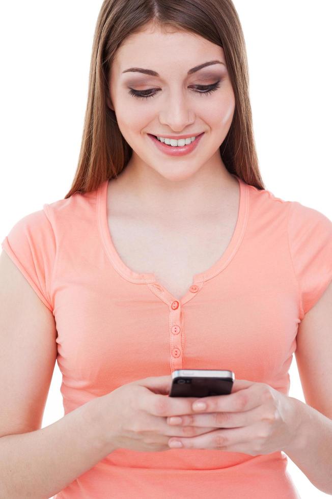 Beauty typing a message. Attractive young woman holding mobile phone and smiling while standing isolated on white photo