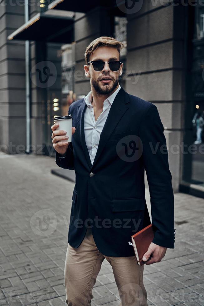 Always in a hurry. Handsome young man in smart casual wear carrying disposable cup while walking through the city street photo