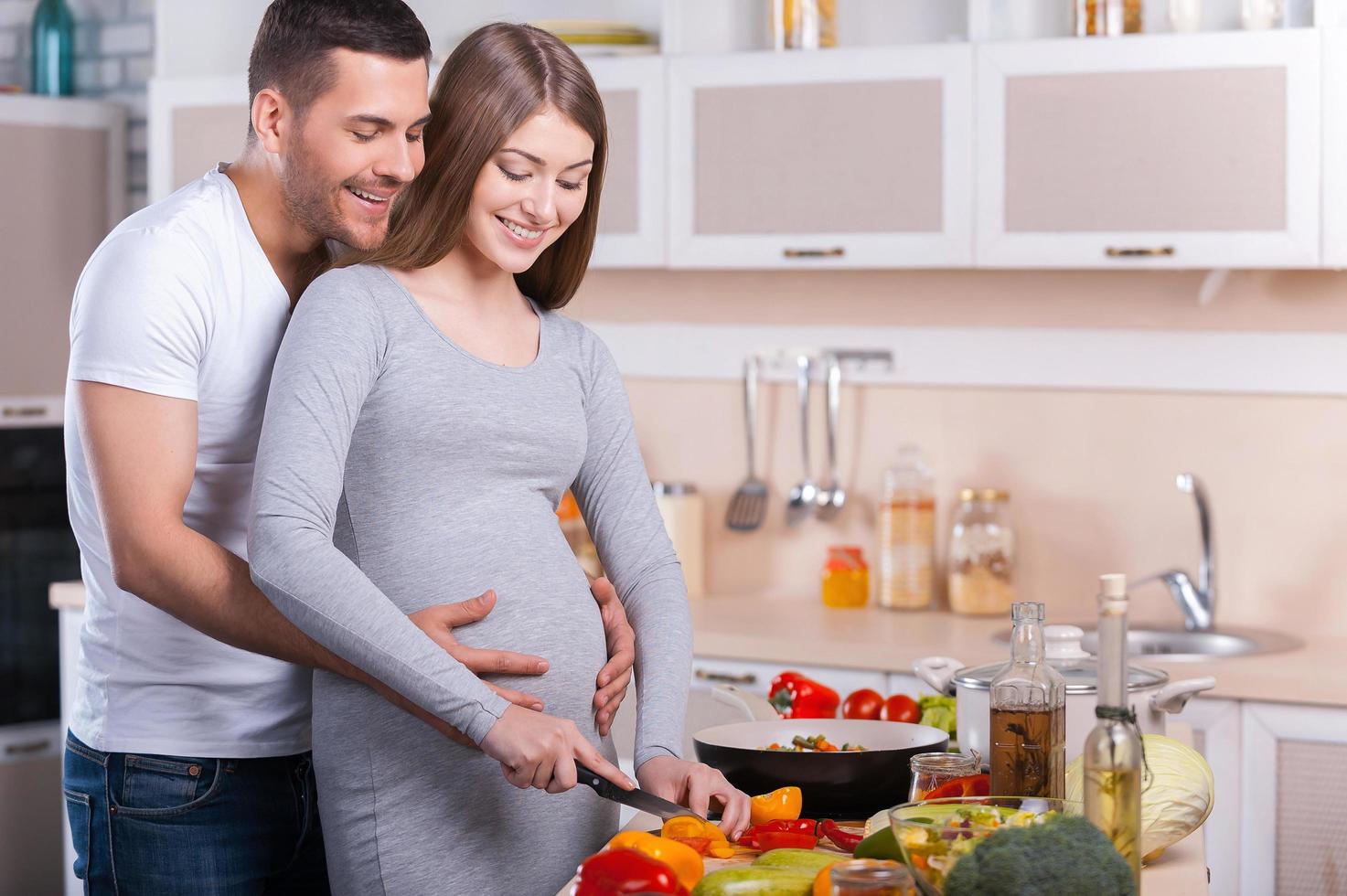 cocinar comida saludable. feliz pareja joven cocinando juntos en la cocina mientras el hombre toca el abdomen de su esposa embarazada foto