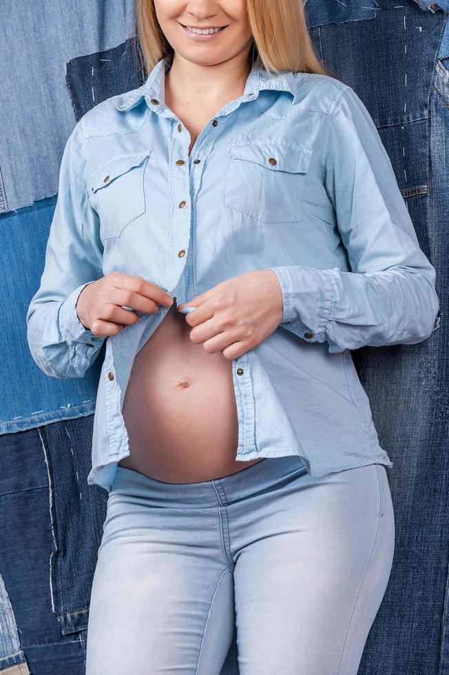 Staying trendy. Cropped image of pregnant woman dressing up her jeans shirt while standing against jeans background photo