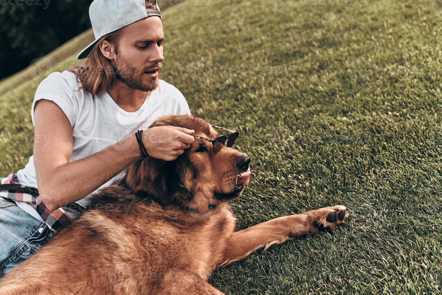 perro con estilo. un joven apuesto que le pone gafas de sol a su perro mientras está acostado en el césped en el parque foto