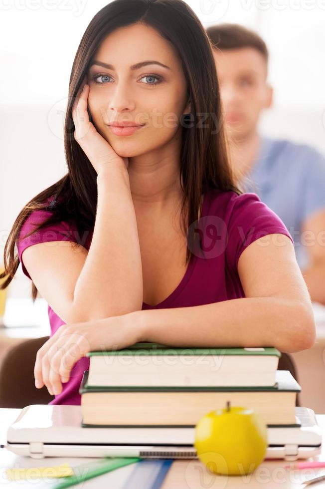 Smart and confident. Beautiful female student sitting at the desk and smiling at camera while young man sitting behind her photo