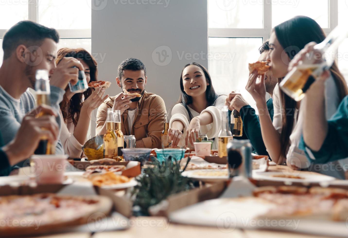 More laugh with friends. Group of young people in casual wear eating pizza and smiling while having a dinner party indoors photo