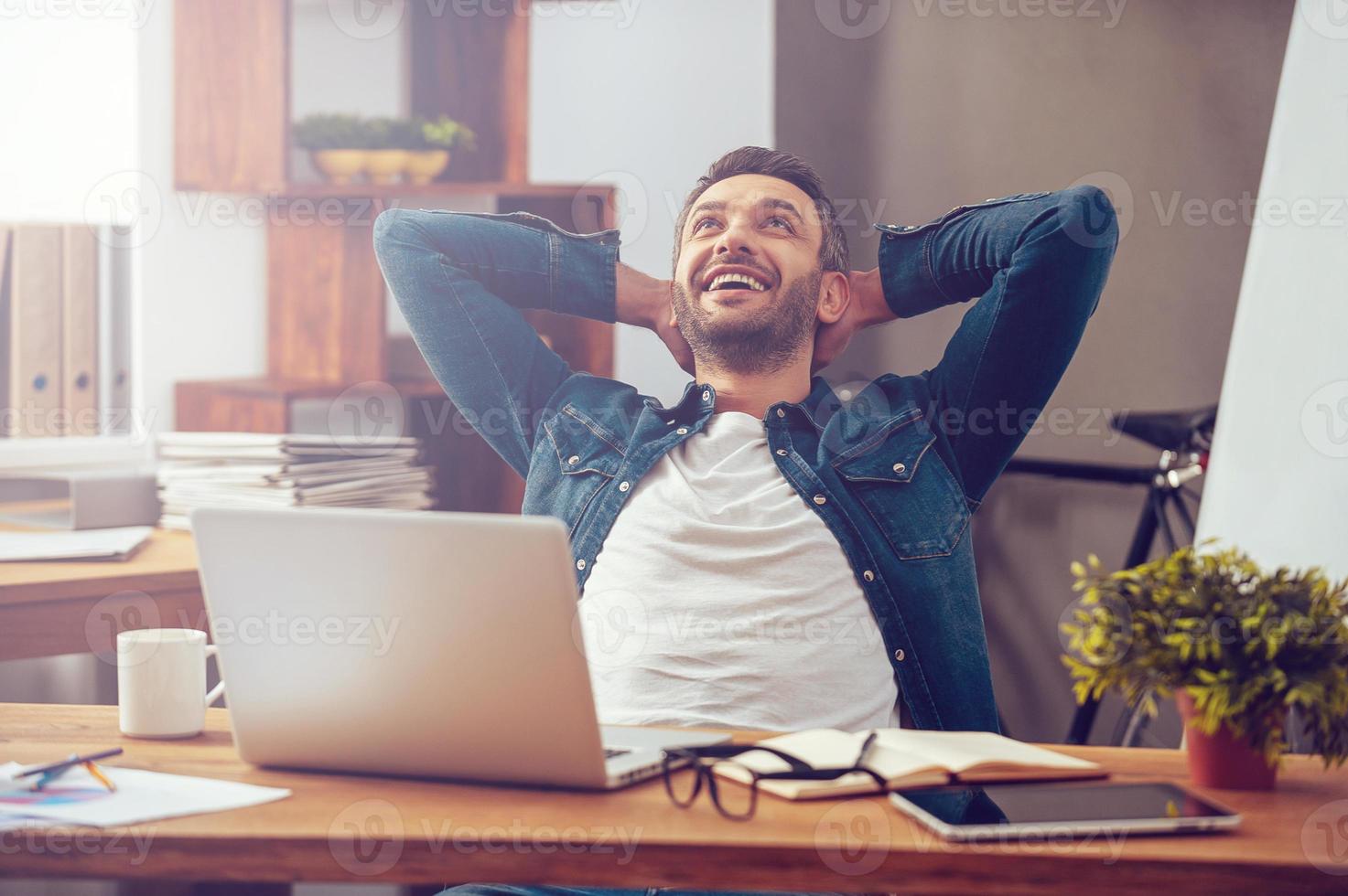 Satisfied with work done. Happy young man working on laptop while sitting at his working place in office photo