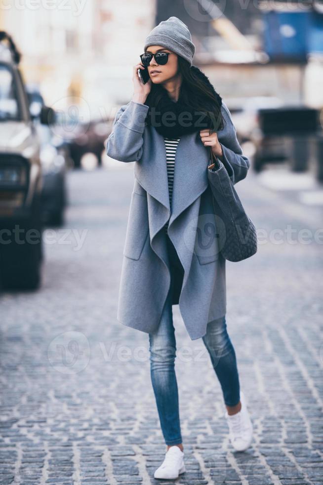 I will be in five minutes. Beautiful young woman in sunglasses talking on mobile phone and looking away while crossing the street photo