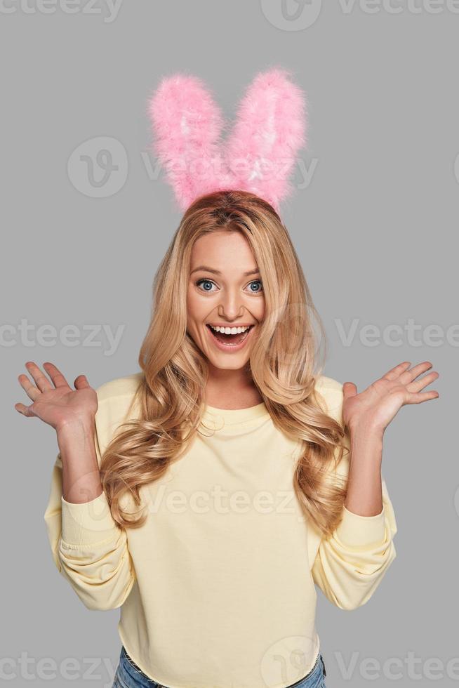 No way Surprised young smiling woman in pink bunny ears gesturing and looking at camera while standing against grey background photo