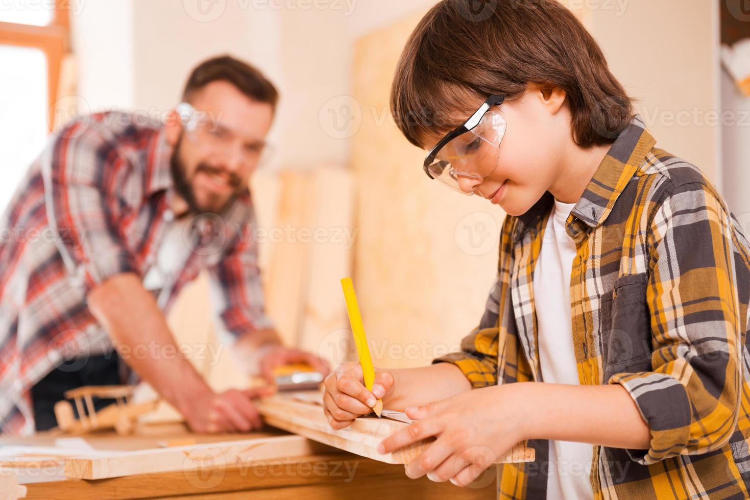 Hardworking is a key to success. Smiling little boymaking measurements on the wooden plank while working with his father in workshop photo