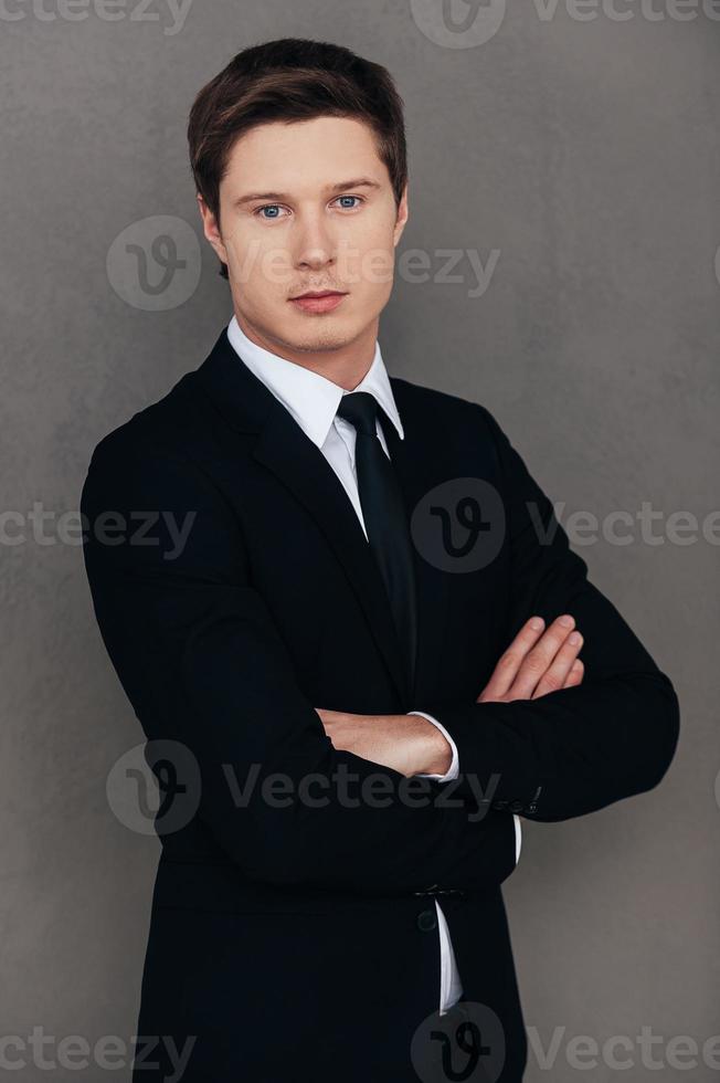 Confident and successful. Confident young man in formalwear keeping arms crossed and looking at camera while standing against grey background photo