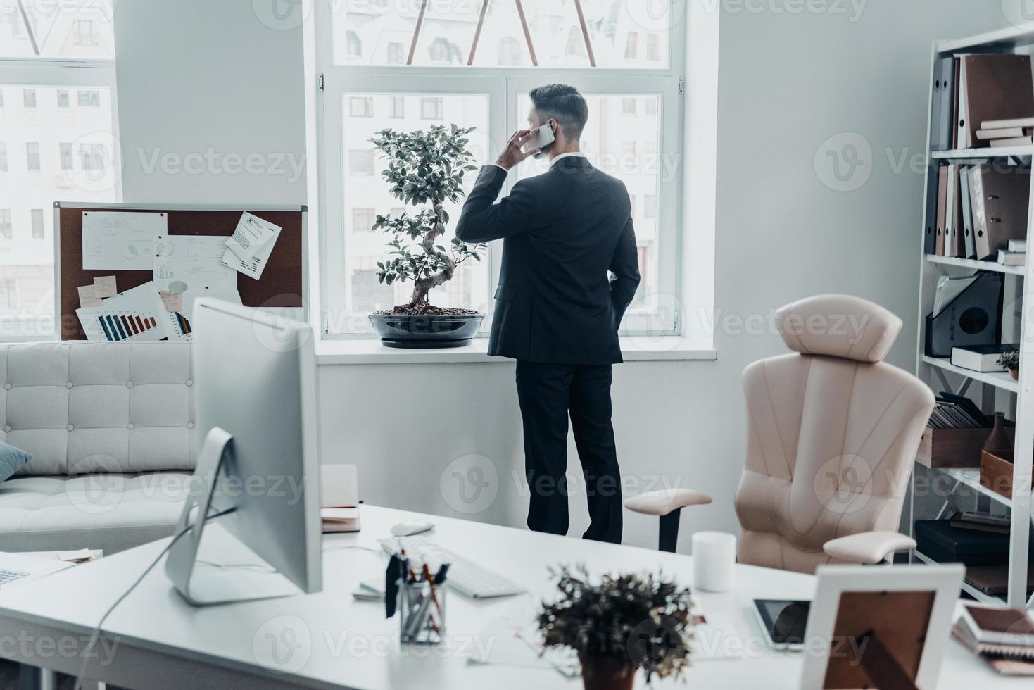 No time for resting. Rear view of young man in full suit talking on the phone and keeping hand in pocket while standing near the window in the office photo