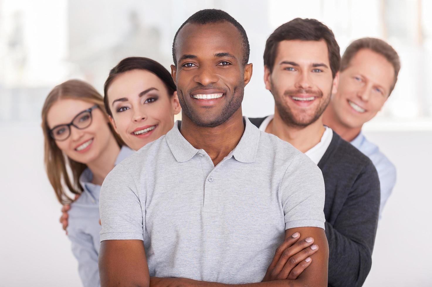 Confident business team. Cheerful young African man keeping arms crossed and smiling while group of people standing behind him in a row and looking at camera photo