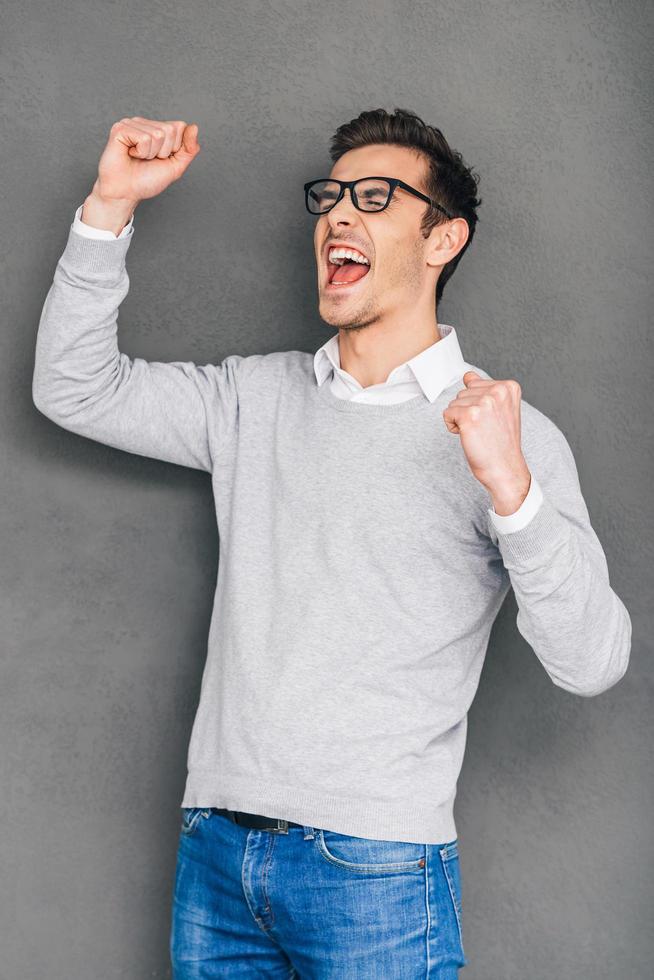 Happy winner. Cheerful young man gesturing and keeping his mouth open while standing against grey background photo