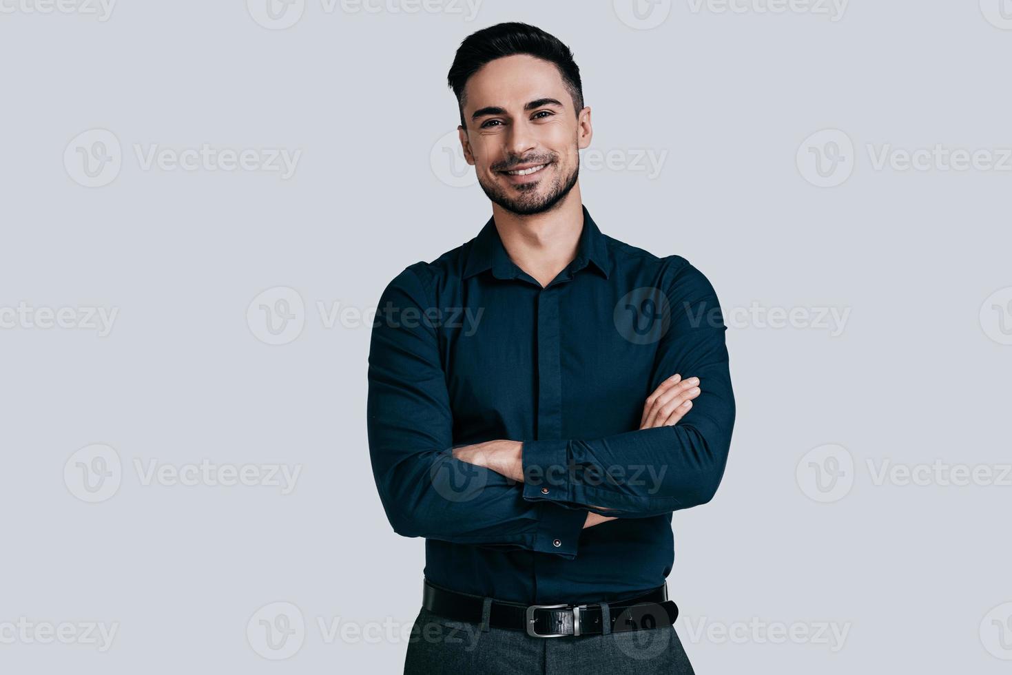 Confident manager. Handsome young man in shirt keeping arms crossed and looking at camera while standing against grey background photo