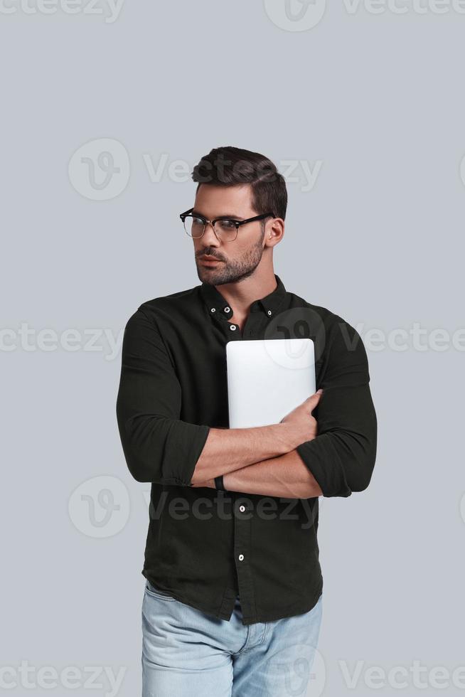 Always online. Good looking young man in eyeglasses holding his digital tablet and looking away while standing against grey background photo